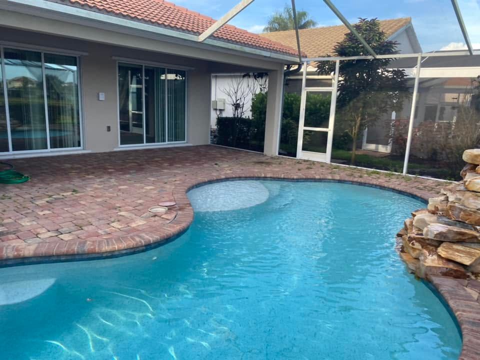 Backyard pool with curved blue water, brick patio, and covered patio beside a house