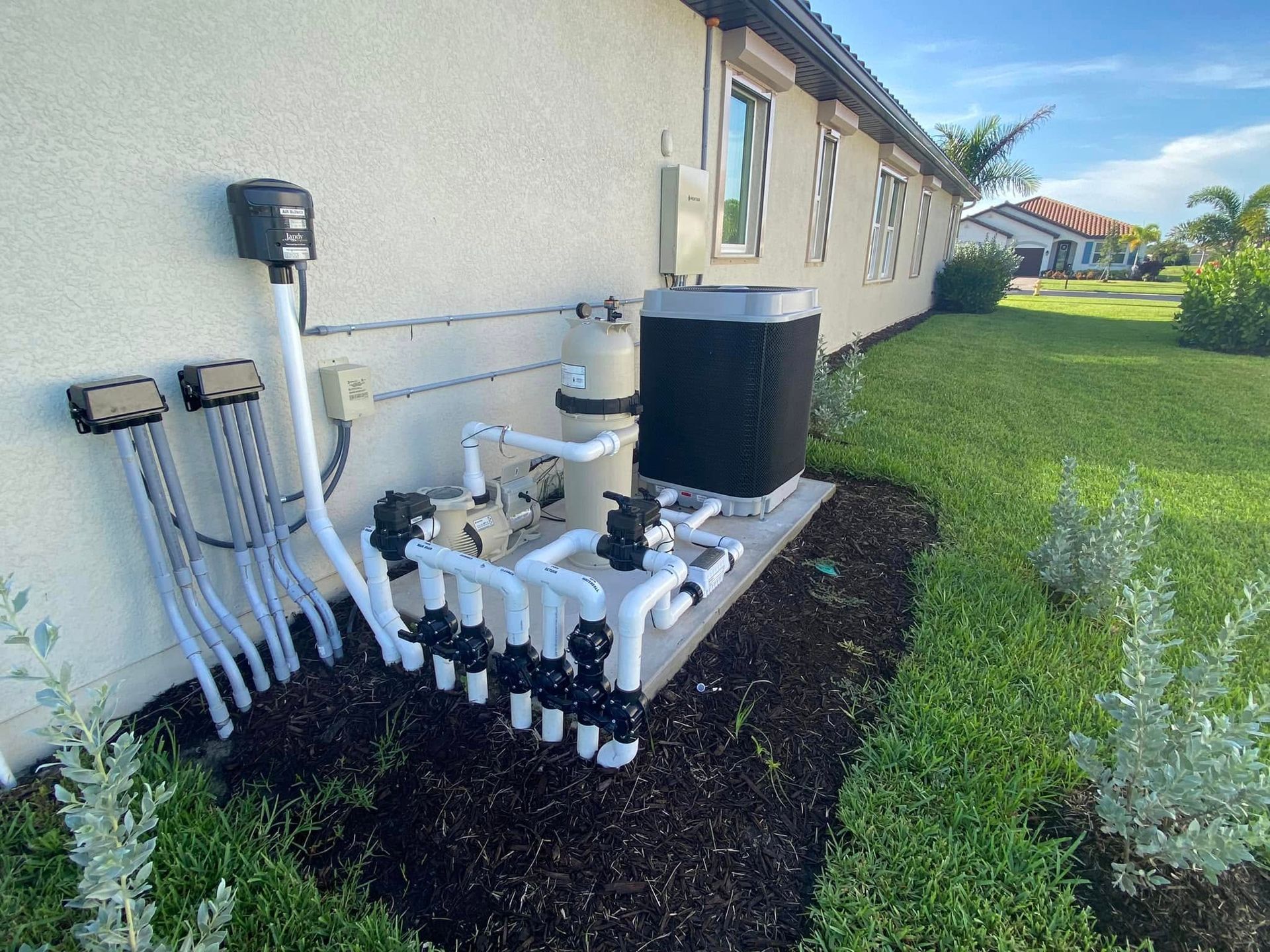 Outdoor pool equipment and plumbing beside a house, with black and white pipes and a water tank.