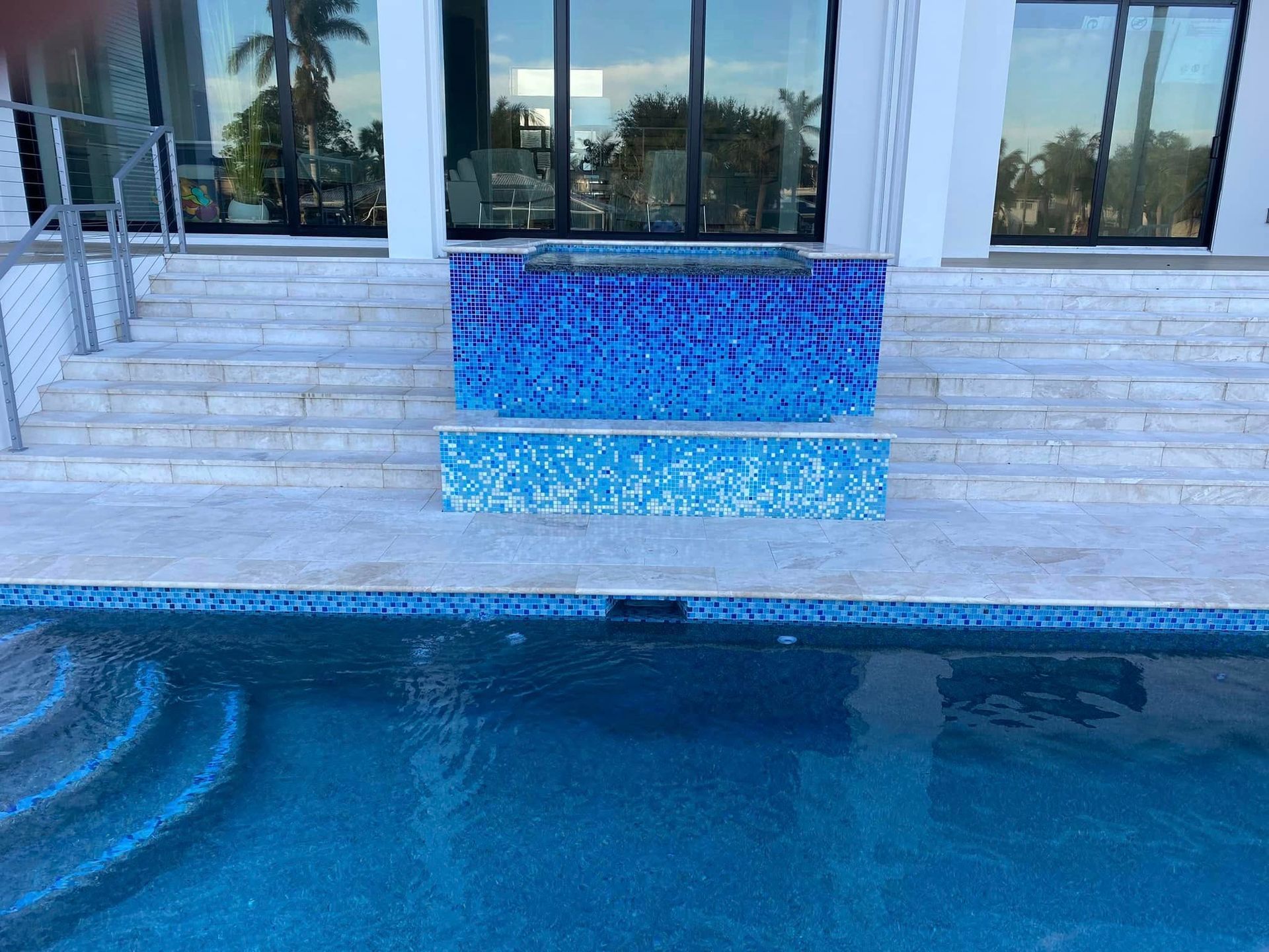 Blue-tiled pool waterfall with steps leading to a house patio and glass doors