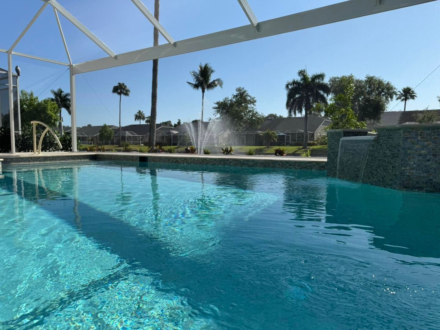 Blue swimming pool with fountain, screened enclosure, palm trees, and bright sunny sky