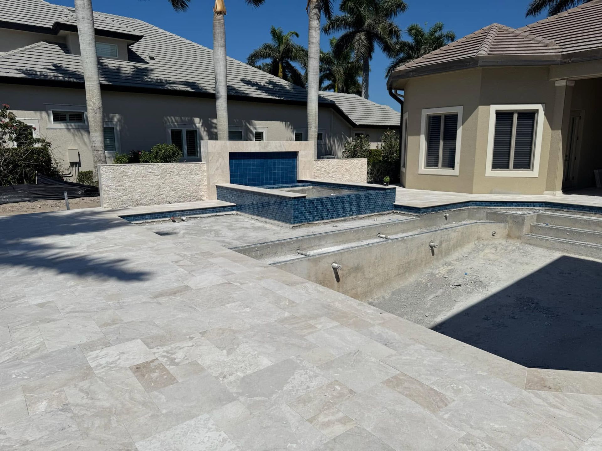Empty backyard pool with blue-tiled spa, stone patio, and beige house under a sunny sky