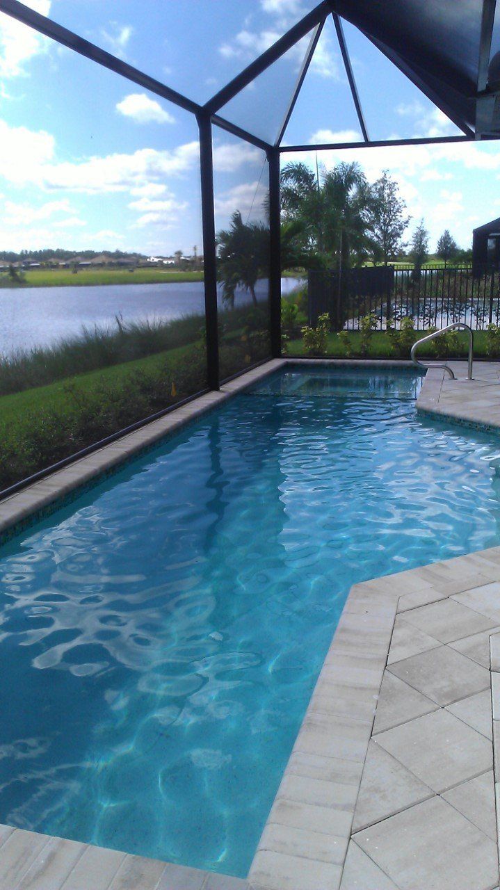 Screened backyard pool beside a lake, with blue water, patio tiles, and a cloudy sky.