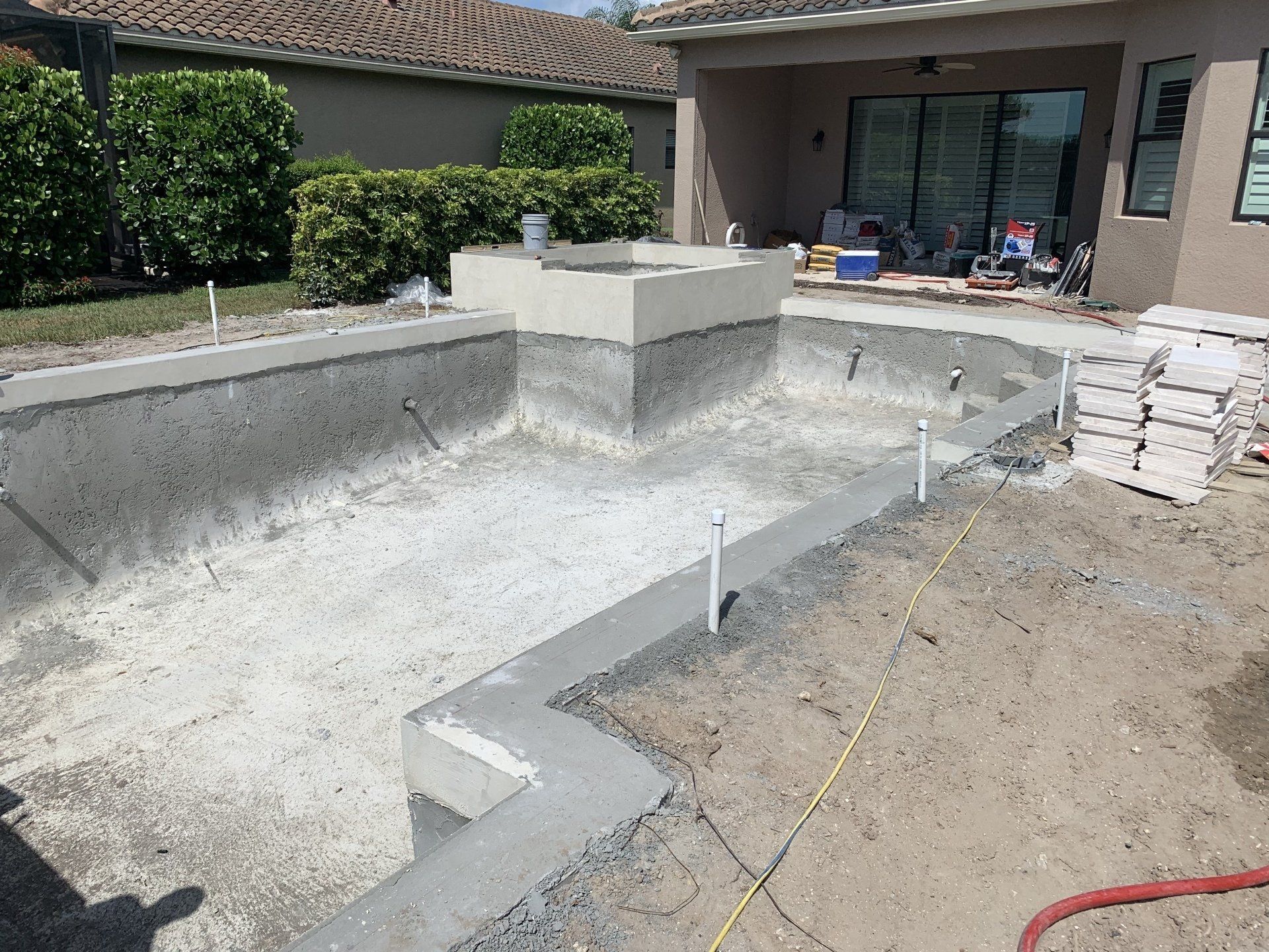 Empty concrete backyard pool under construction beside a house, with stacked tiles and work materials nearby