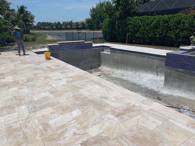 Empty outdoor pool area with light stone decking, a worker, and a yellow bucket under sunny skies