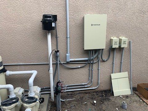 Wall-mounted utility equipment with pipes, electrical boxes, and a beige panel on a textured exterior wall.