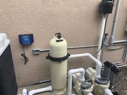 Pool equipment with beige filter tank, pipes, and pumps against a stucco wall.