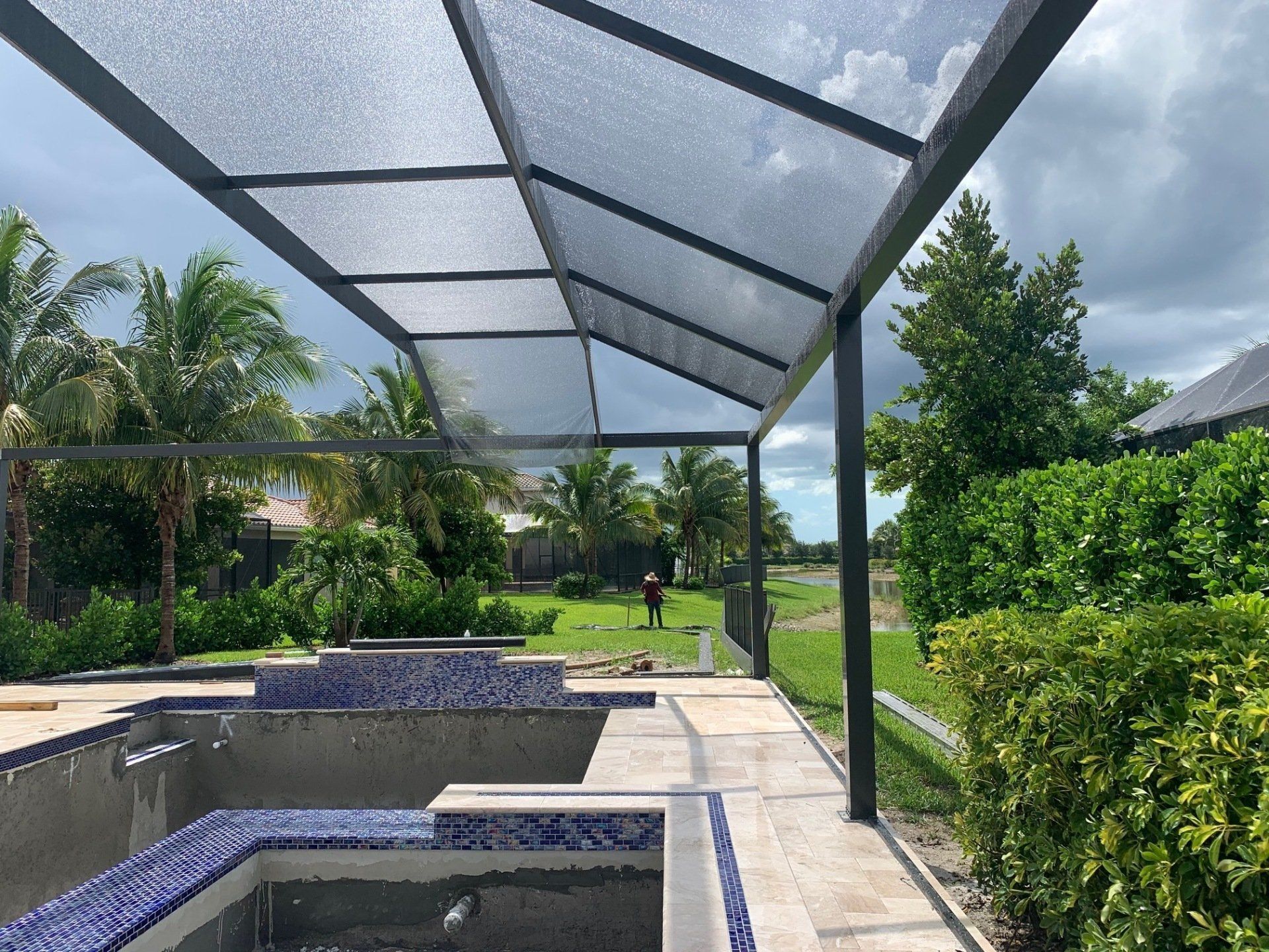 Covered pool patio with blue-tiled spa, screened roof, and lush green yard under cloudy sky