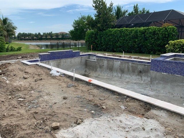 Empty in-ground pool under construction beside a house, with dirt foreground and blue tile edging.