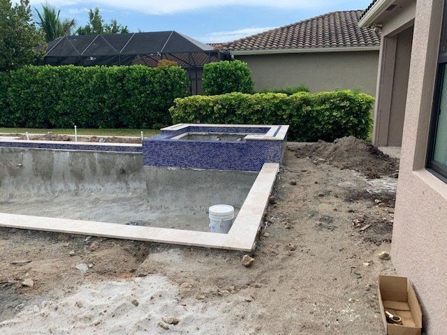 Empty backyard pool beside a house, with unfinished dirt landscaping and a tiled spa under construction