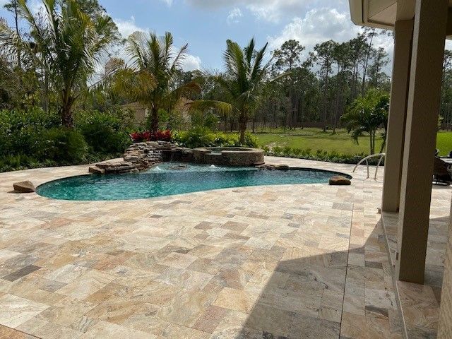 Stone patio with curved turquoise pool, tropical plants, and palm trees beside a covered terrace