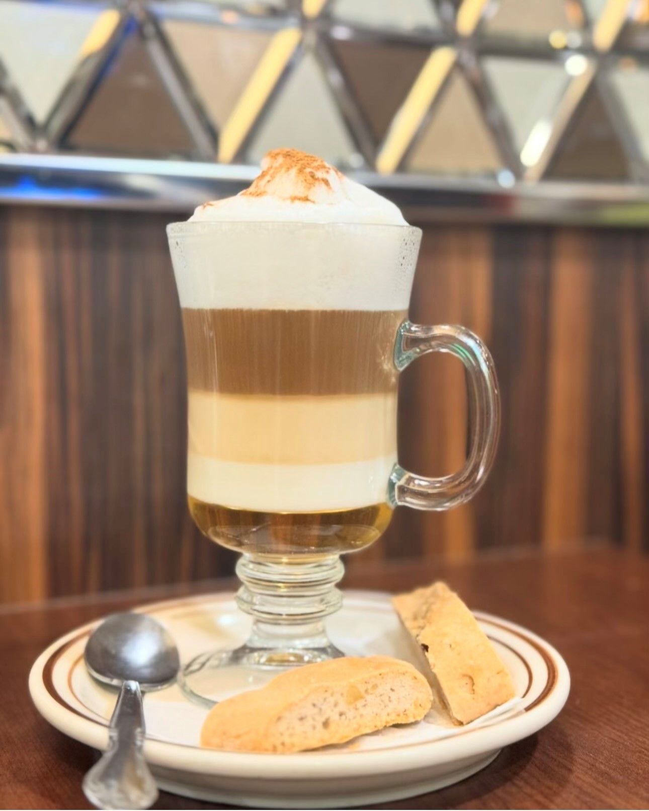 A cup of coffee with whipped cream and a cookie on a plate with a spoon