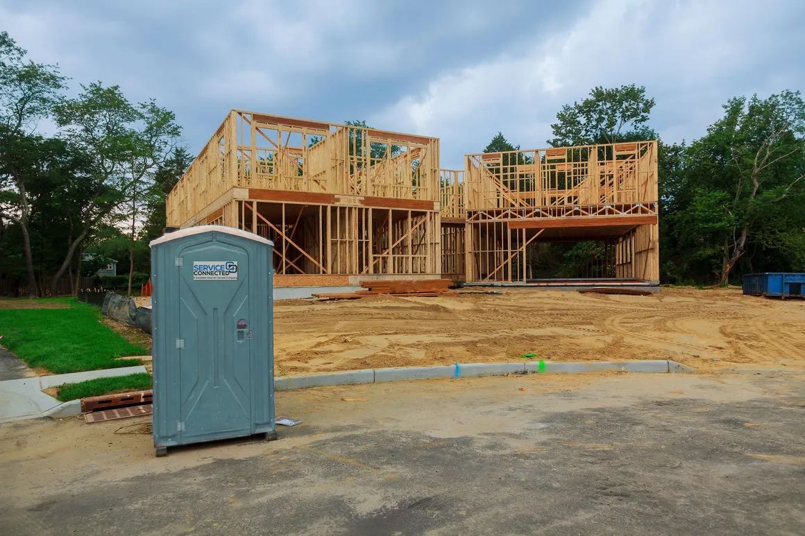 A teal colored portable toilet in front of a partially constructed two-story wooden framed house