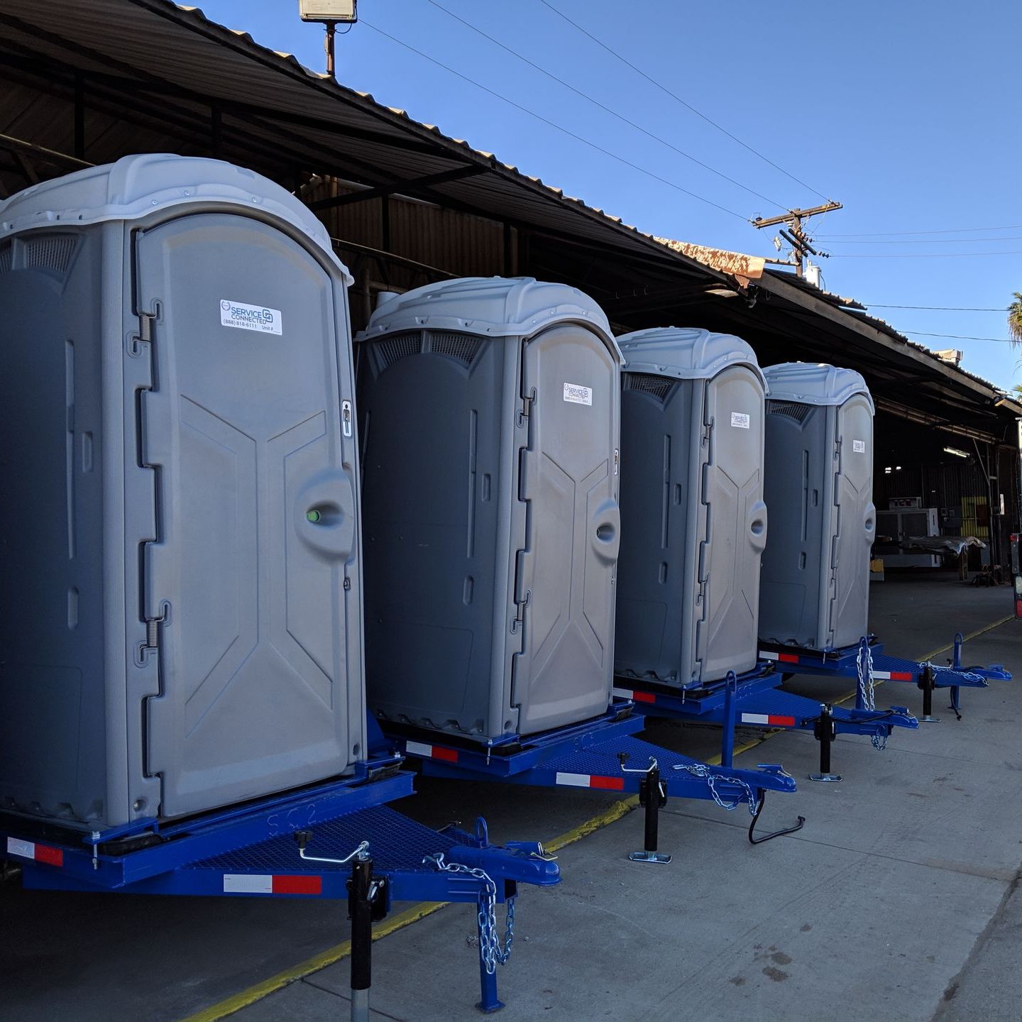 Four gray portable toilets lined up on blue trailers under a shelter.
