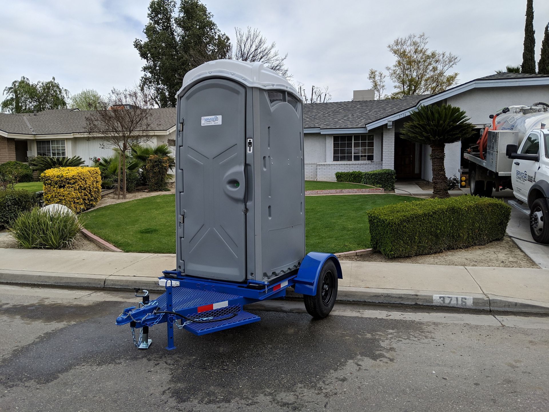 A gray portable toilet on a blue trailer parked on a residential street.