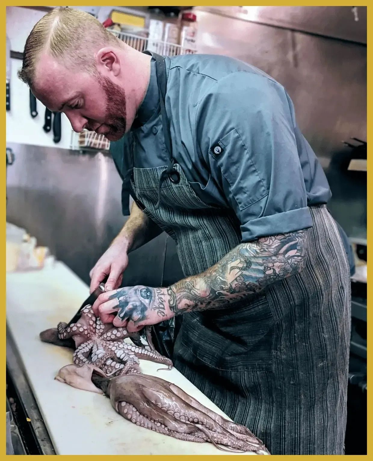 Chef butchering a squid in a kitchen, with tattooed arms and a black apron.