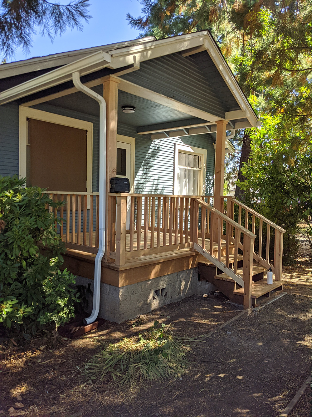 Small porch entrance with wooden railing, steps, and a downspout on a brown house in a wooded yard