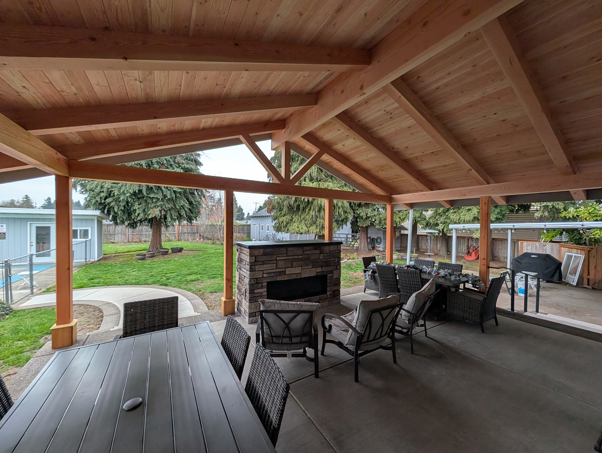 Covered outdoor patio with wooden roof, fireplace, and dining table with chairs.