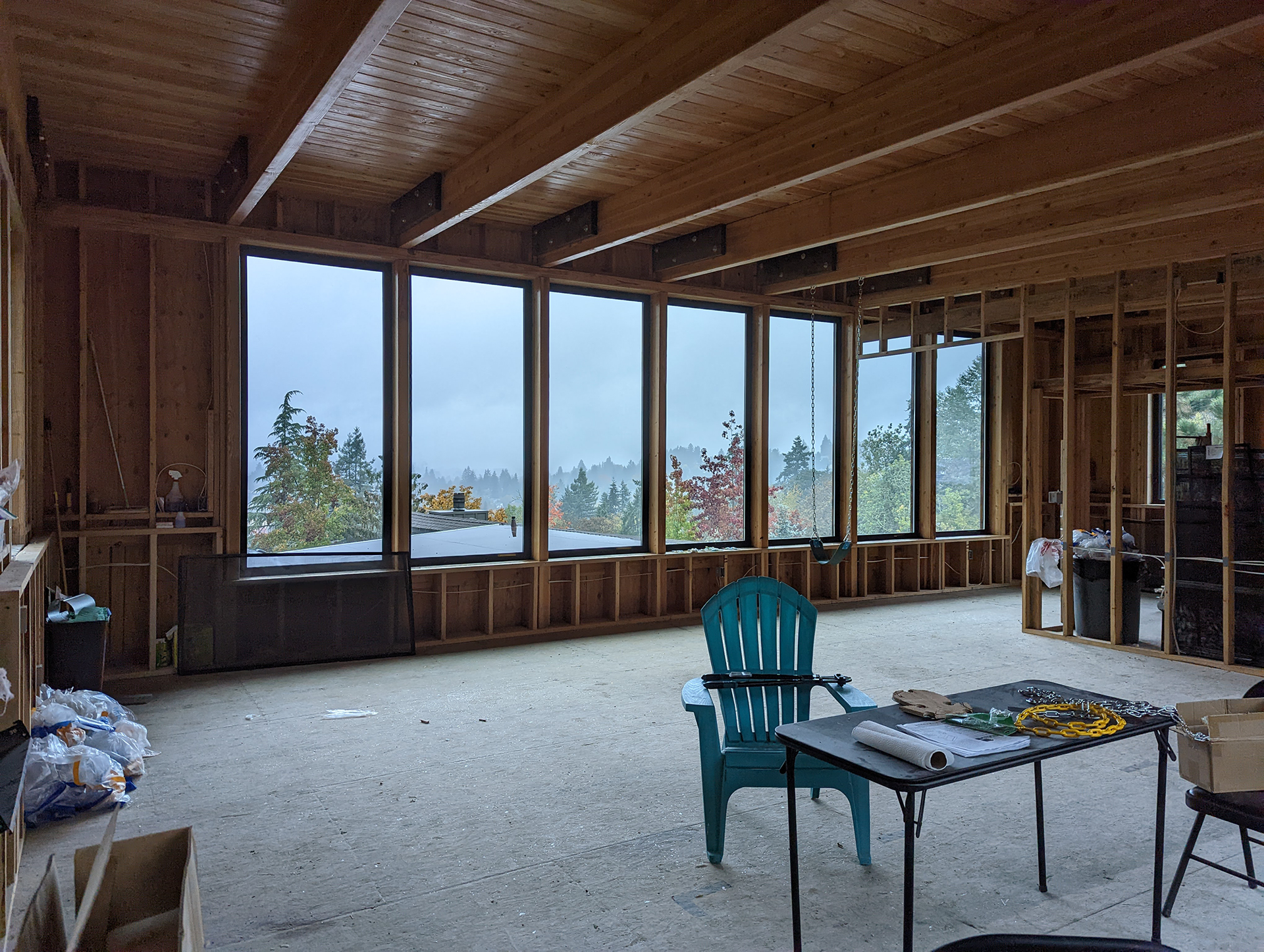 Unfinished room with large windows, exposed wood framing, and a teal chair beside a small table.