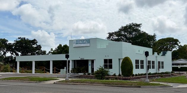 Low white building with columns and large windows beside a road under a cloudy sky