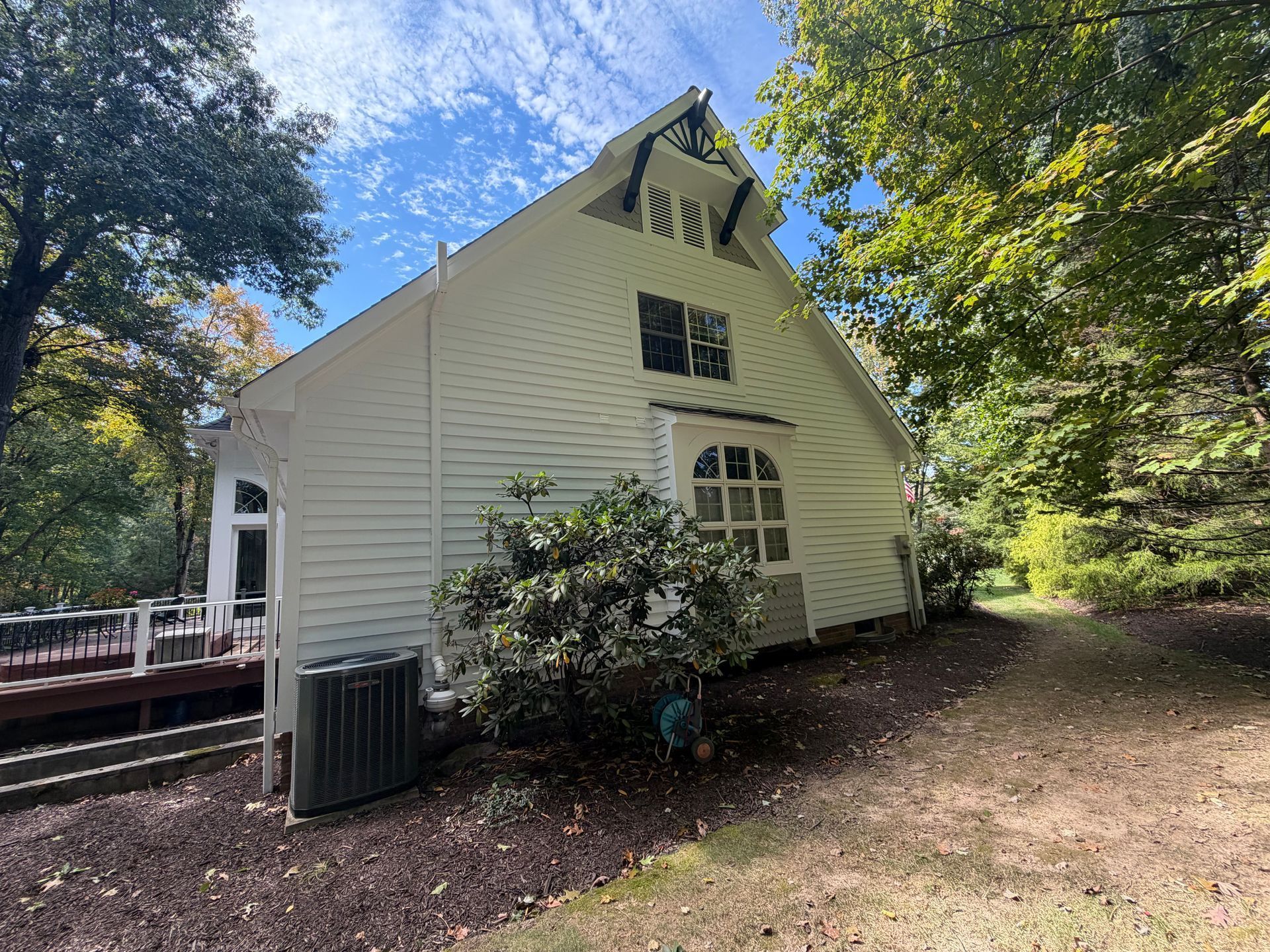 White house with steep roof and small porch, surrounded by trees and mulch in a wooded yard