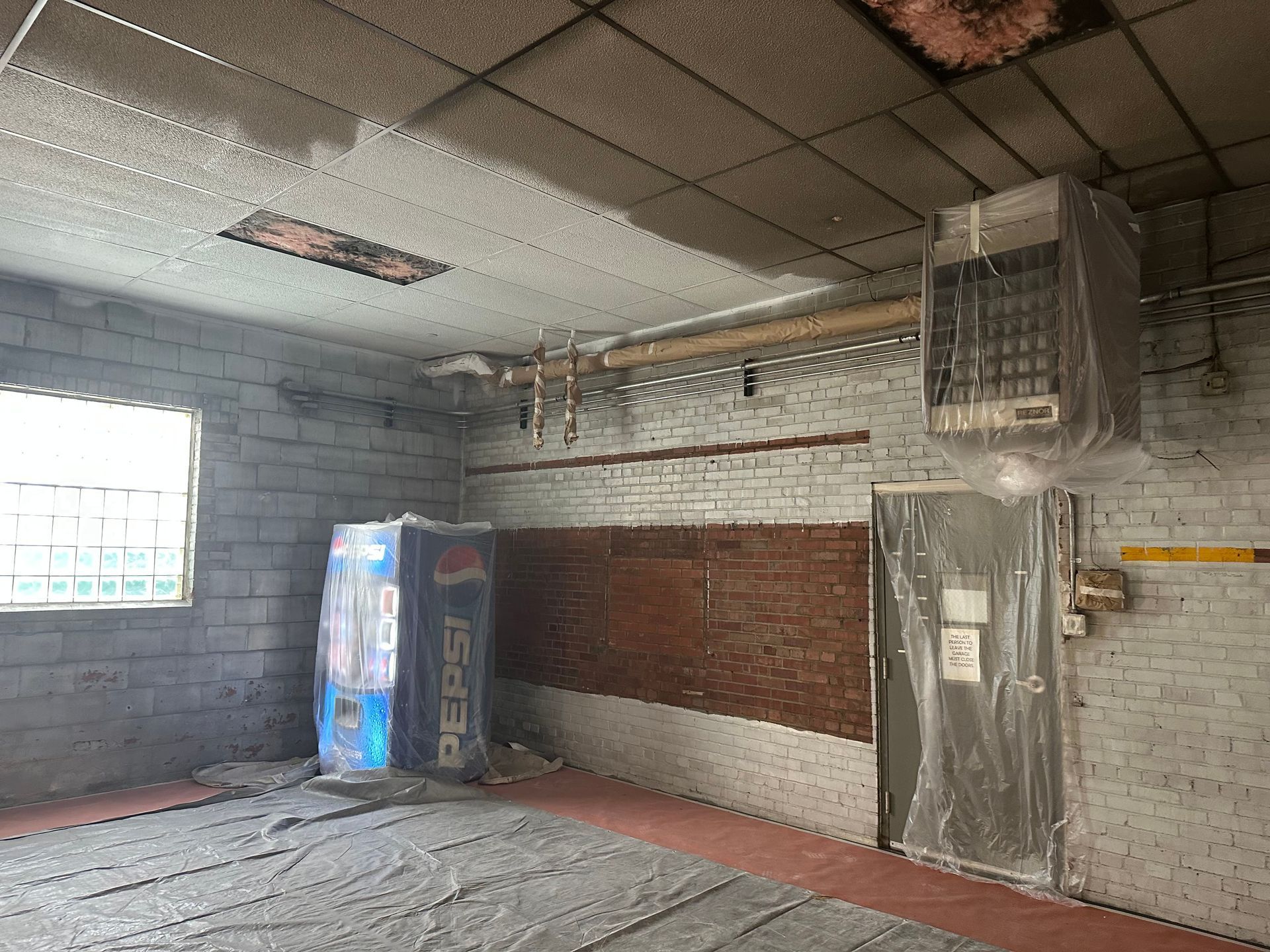 A room with a Pepsi machine, exposed brick, a heater, and a plastic-covered door.