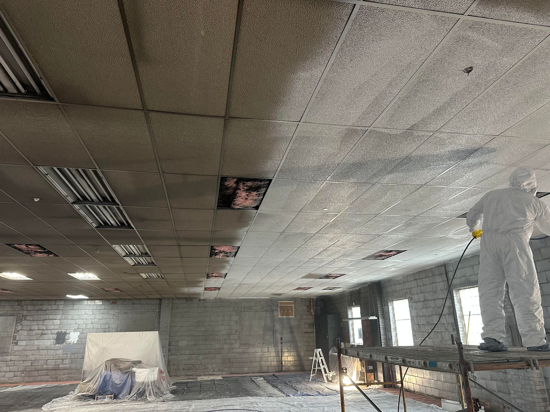 Worker in protective suit sprays ceiling, partially covered with insulation. Interior construction site.