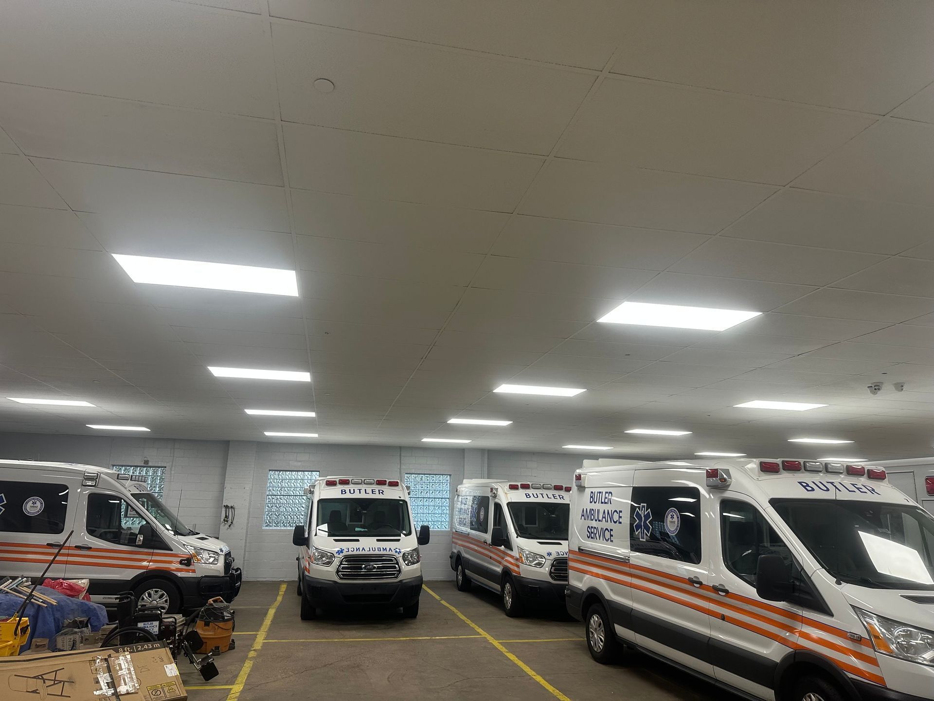 Ambulances parked indoors under fluorescent lights. Several white vehicles with orange stripes in a large garage.