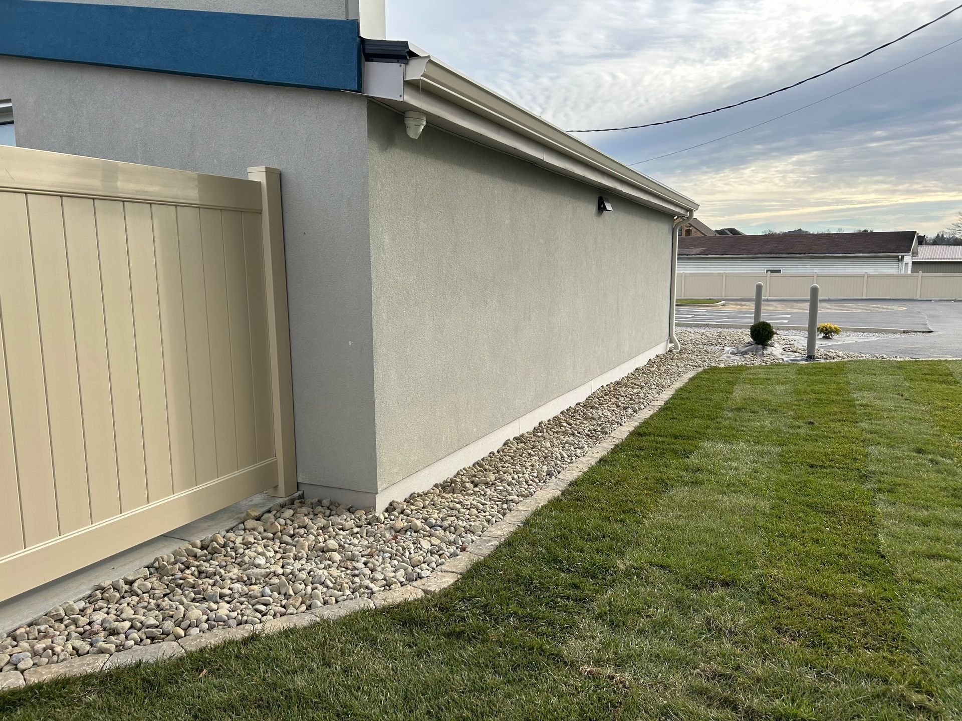 Tan fence borders a building with stucco exterior. A bed of gravel separates building and green lawn.