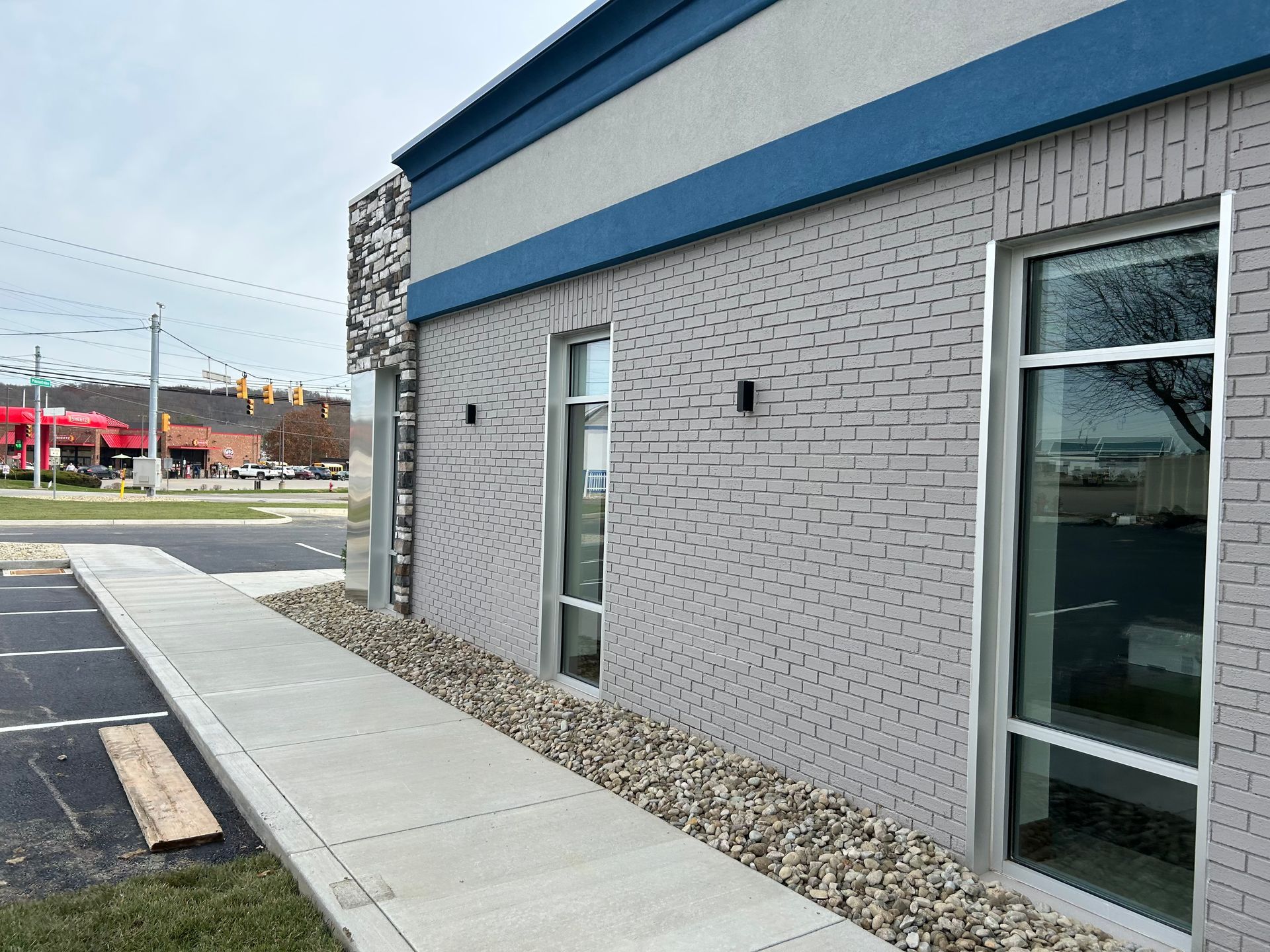 Exterior of a building with painted gray brick, blue trim, and sidewalk. Large windows and a rock bed are visible.