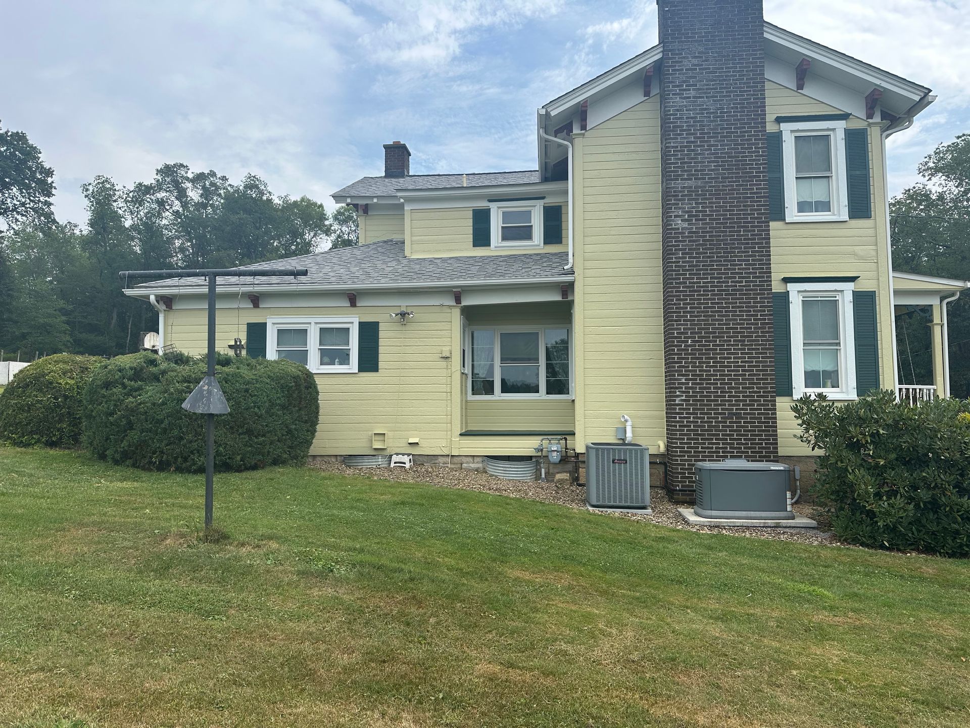 Yellow two-story house with a large brick chimney, green shutters, and a grassy yard on an overcast day.