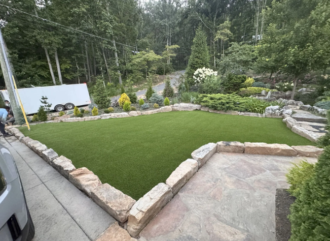 A manicured lawn bordered by stone, adjacent to a driveway and landscaping, with a forest in the background.