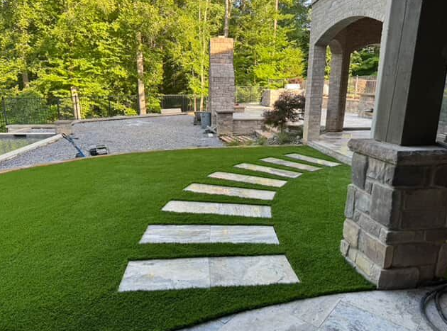 A stone pathway on green turf leads to an outdoor patio with a wooded backdrop.