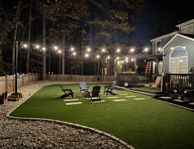 Backyard with string lights, fire pit, and chairs on a green lawn at night.