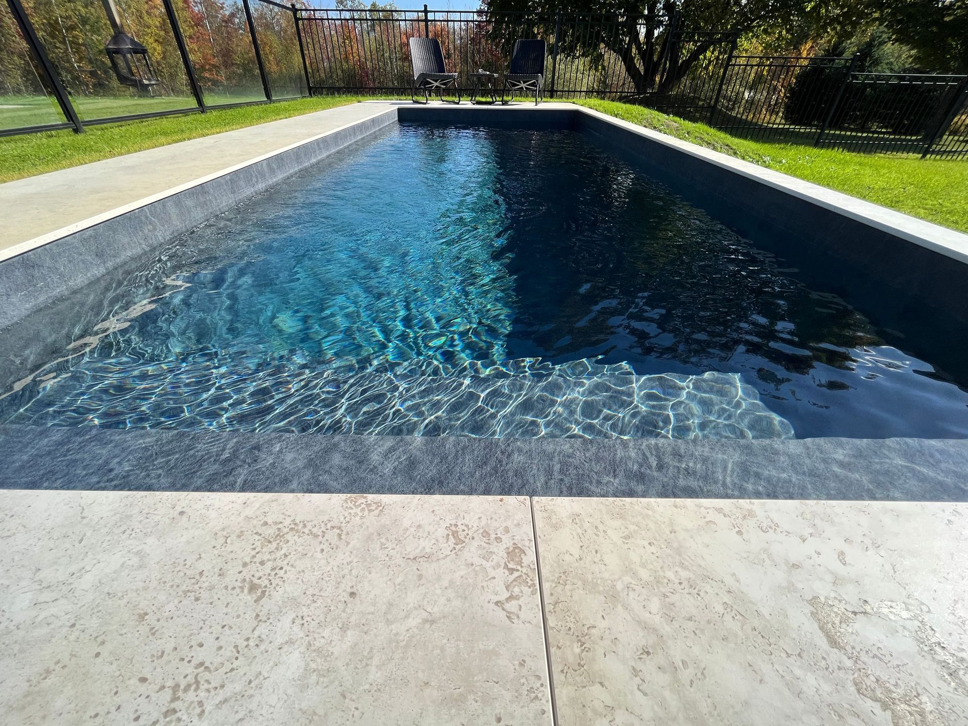Rectangular blue swimming pool with sunlit water and a paved deck, bordered by grass and a fence.
