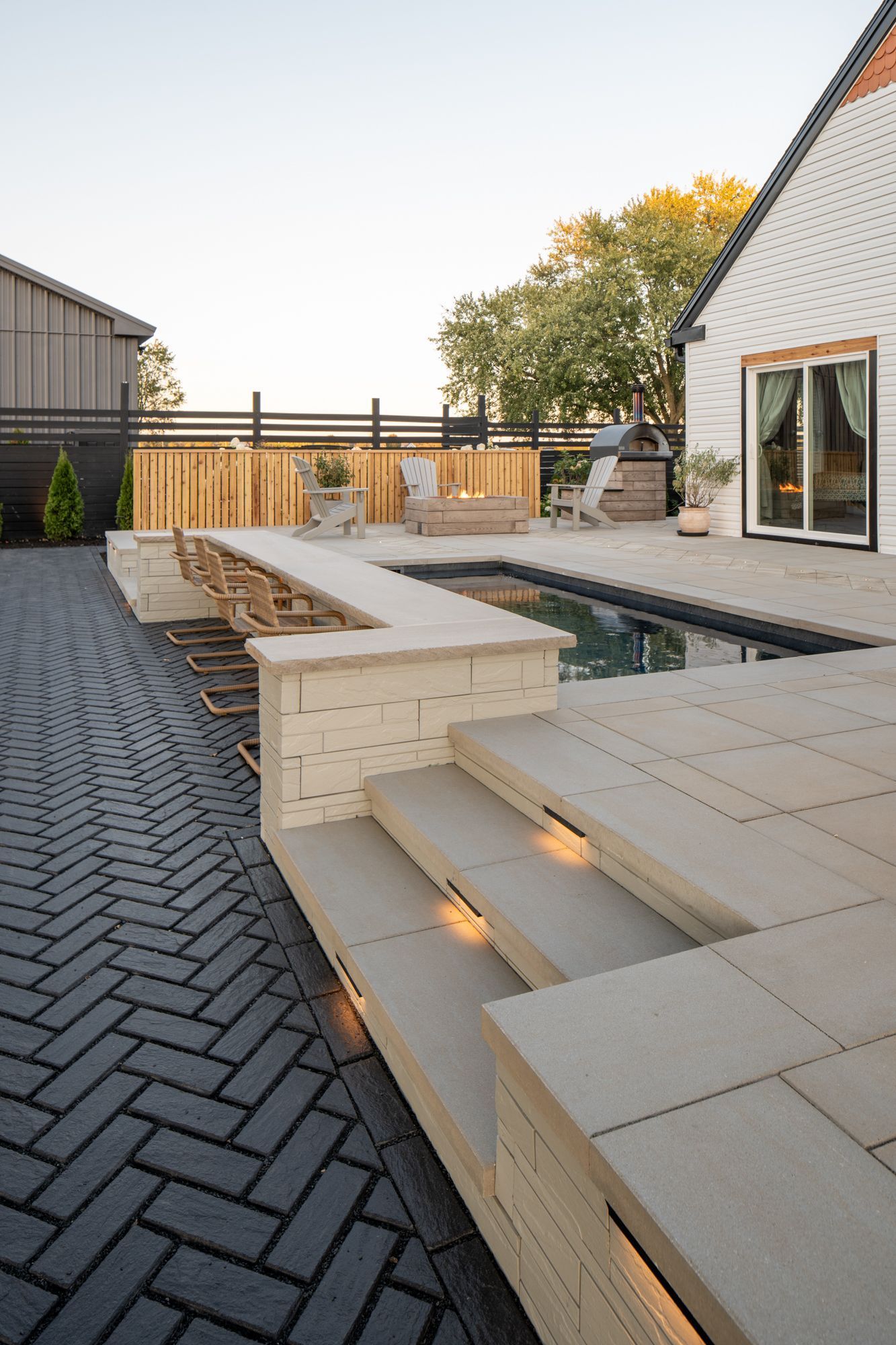 Modern backyard patio with light wood benches, dark pavers, and a rectangular pool beside a house