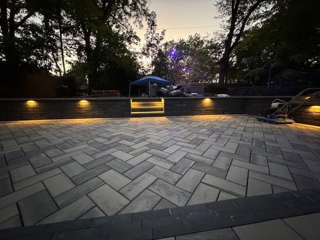Brick patio with illuminated retaining wall, steps, and trees at dusk.