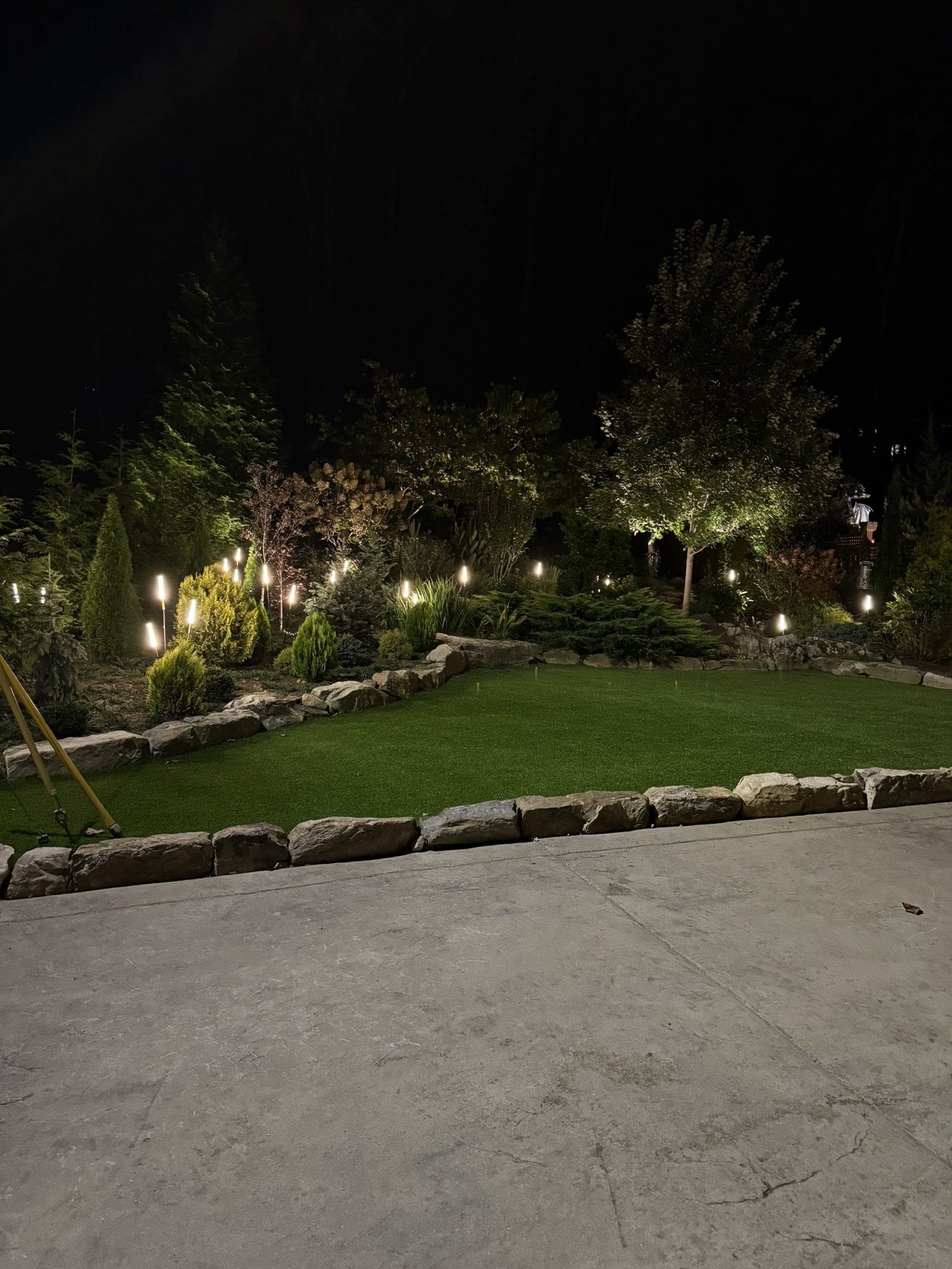 Nighttime garden with string lights illuminating grass, trees, and stone borders.