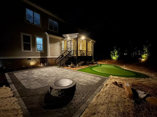 Night view of a backyard with a patio, fire pit, putting green, and house with exterior lighting.