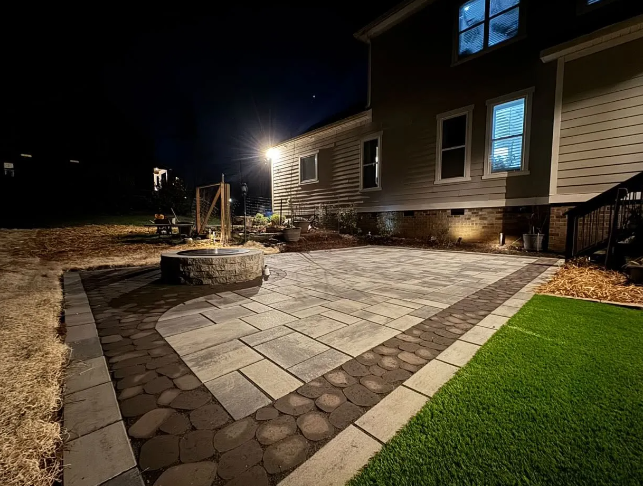 Nighttime view of a backyard patio with pavers, fire pit, and house with lit windows.