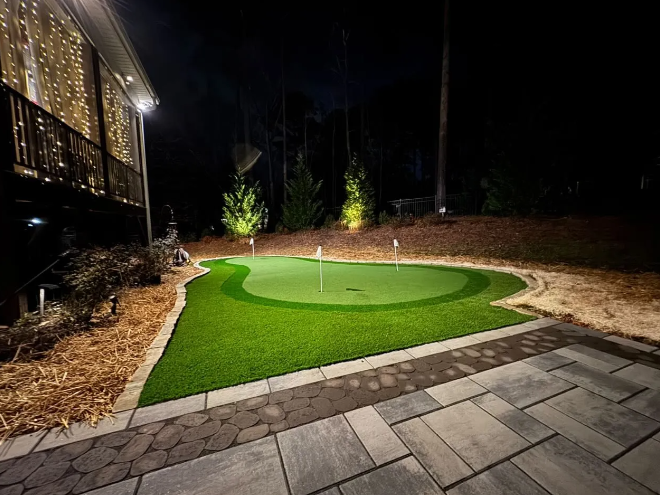 A backyard putting green at night with landscape lighting and a paved patio.