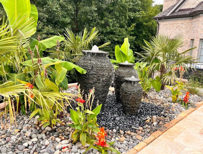 Water fountain feature with three dark stone urns, surrounded by tropical plants and black river rocks.