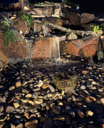 Small waterfall flowing over reddish rocks into a bed of pebbles, illuminated at night.
