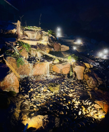 Nighttime view of illuminated tiered waterfall feature with rock formations and small lights.