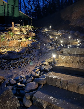 Stone steps illuminated by lights ascend a hillside, alongside a rocky stream.