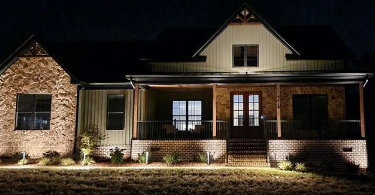 A house at night, lit by spotlights. Brown brick and siding, a covered porch, and a dark sky.