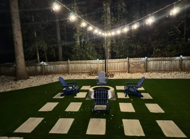 Backyard at night with string lights, fire pit, blue chairs, and stone pavers on green turf.