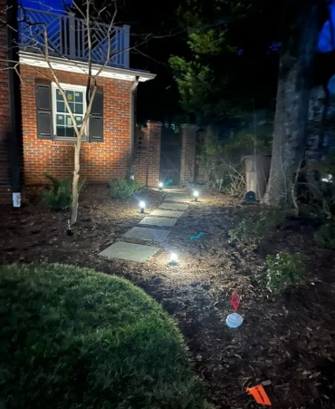 Lit stone pathway leading through a yard at night, with brick building and pillars.