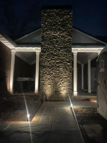 Stone chimney lit at night; walkway with lights leads to a covered entrance with pillars.