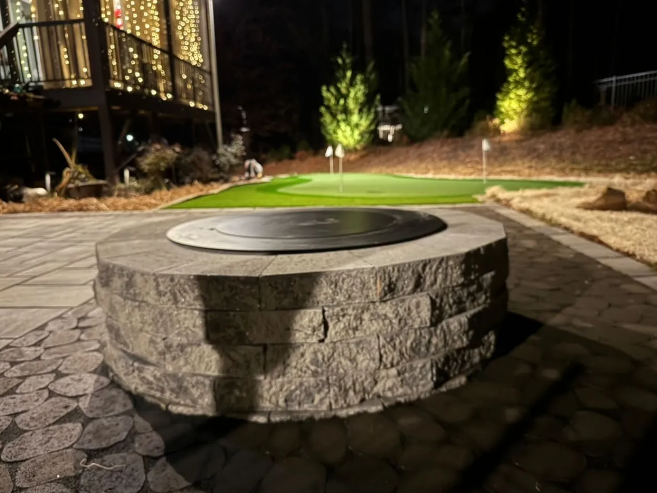Stone fire pit on a patio, with a putting green in the background, lit at night.