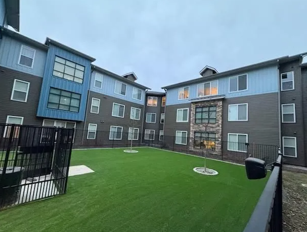 Apartment complex exterior with a courtyard and turf lawn, gray and blue siding, cloudy sky.
